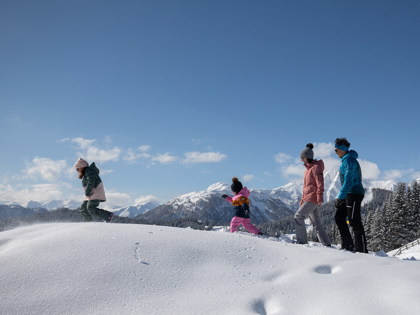 Familie wandert durch unberührte Schneelandschaft mit Ausblick auf die Alpen und verschneite Wälder im Hintergrund.