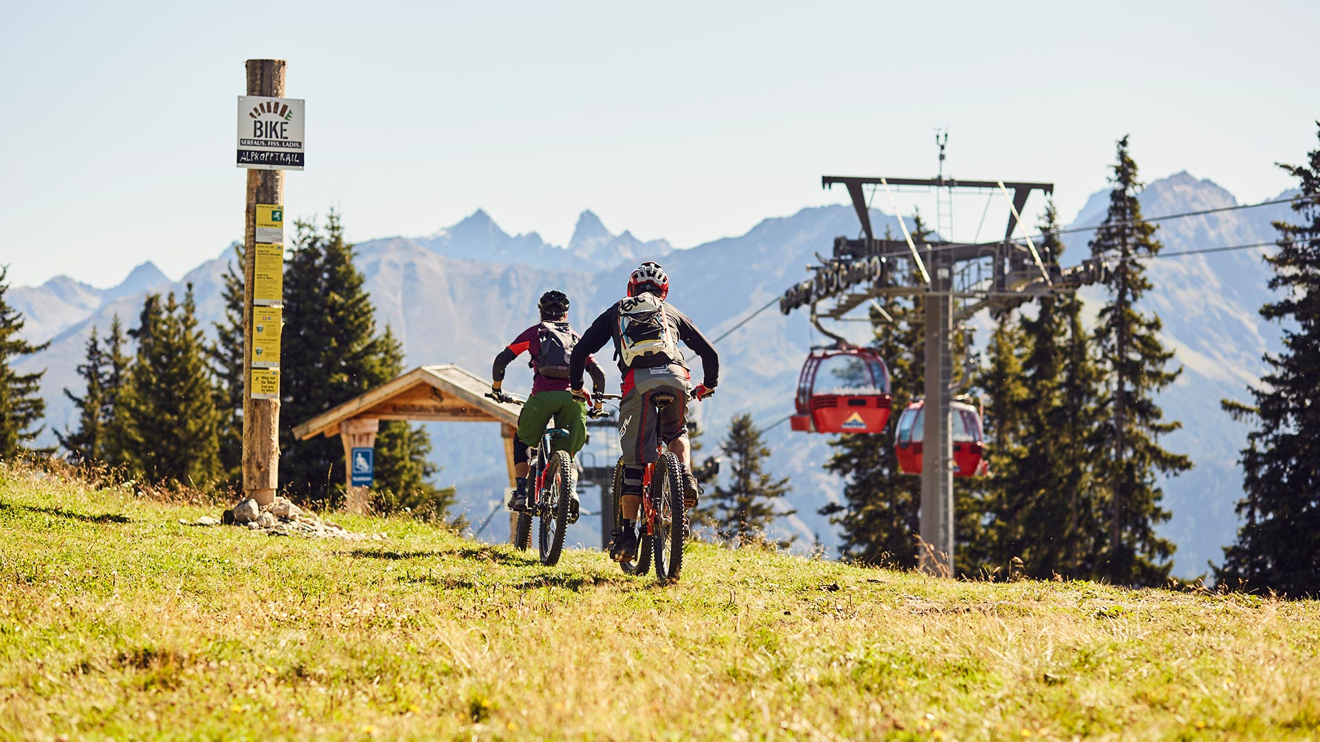Zwei Mountainbiker fahren über eine Almwiese Richtung Sessellift, mit Blick auf die Berge im Hintergrund.