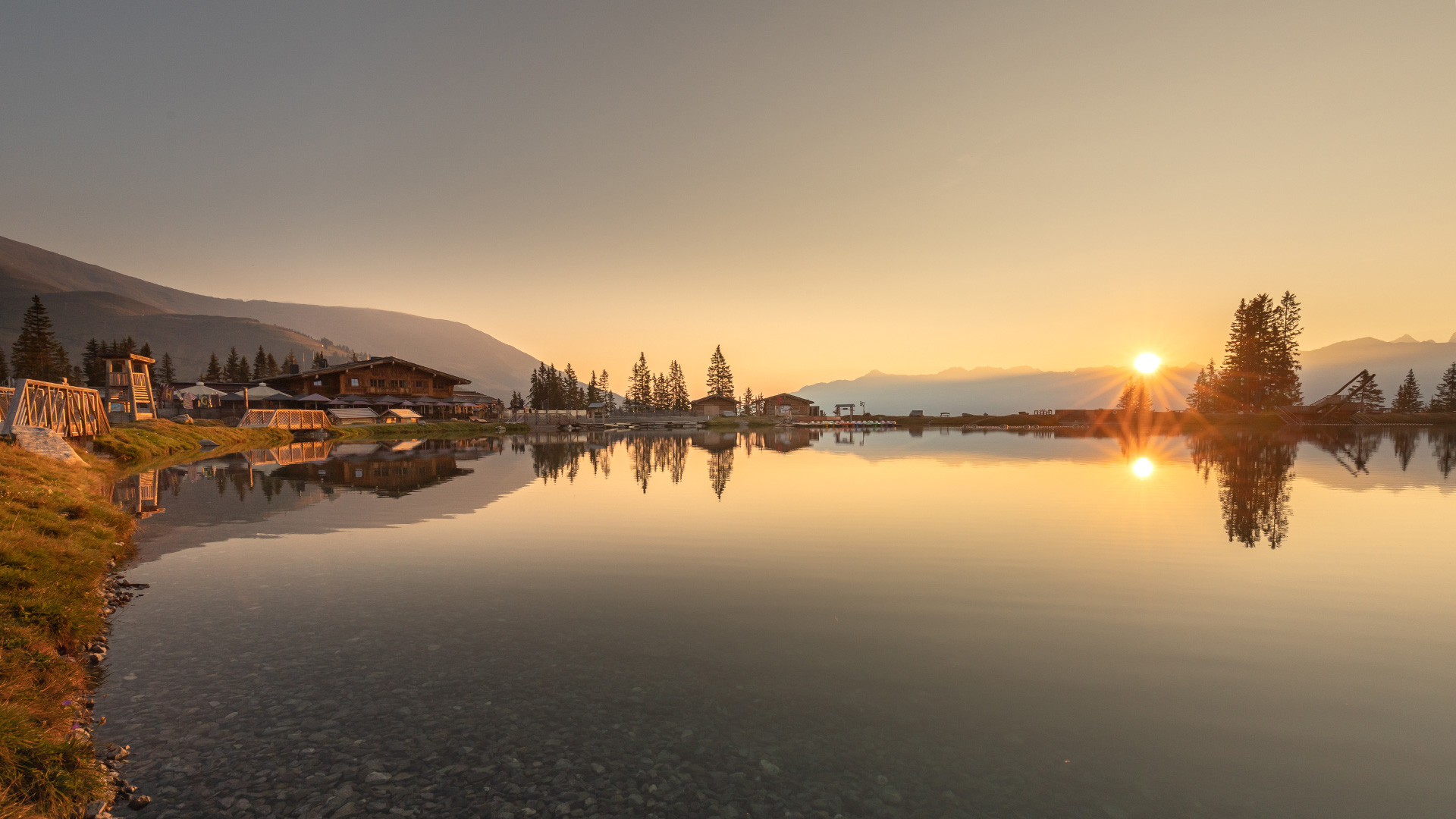 Sonnenaufgang spiegelt sich im ruhigen Bergsee neben einem Chaletdorf mit Wald und Bergen im Hintergrund.