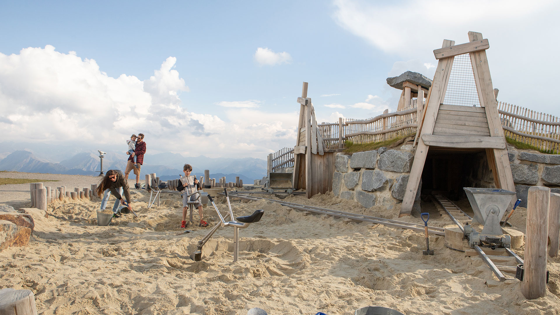 Kinder spielen in einem großen Abenteuersandspielplatz mit Holzburg und Tunnel auf einem Plateau mit Bergpanorama.