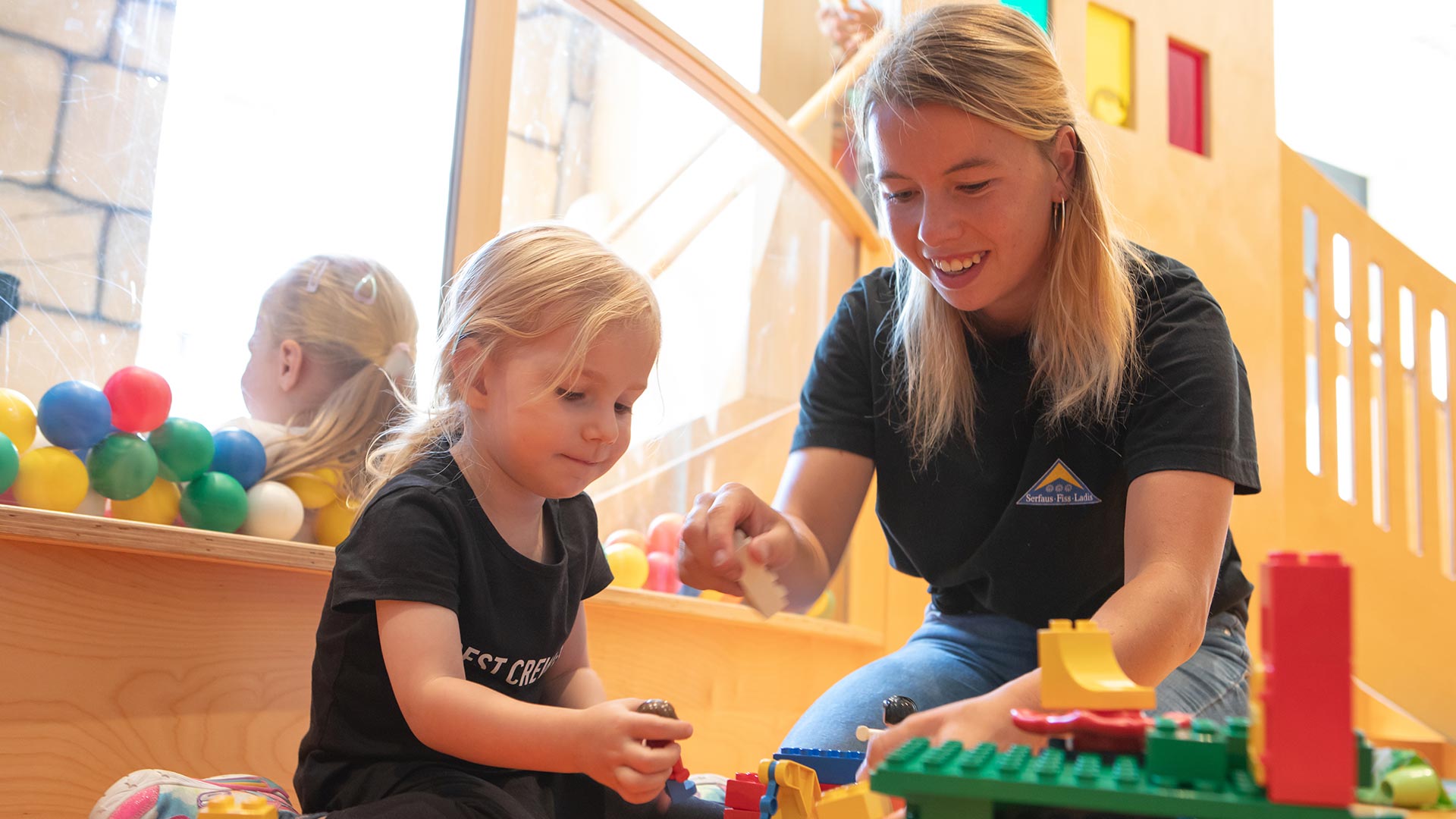 Kleines Mädchen spielt mit einer Betreuerin in einer bunten Indoor-Kinderecke mit Bausteinen und Bällebad im Hintergrund.
