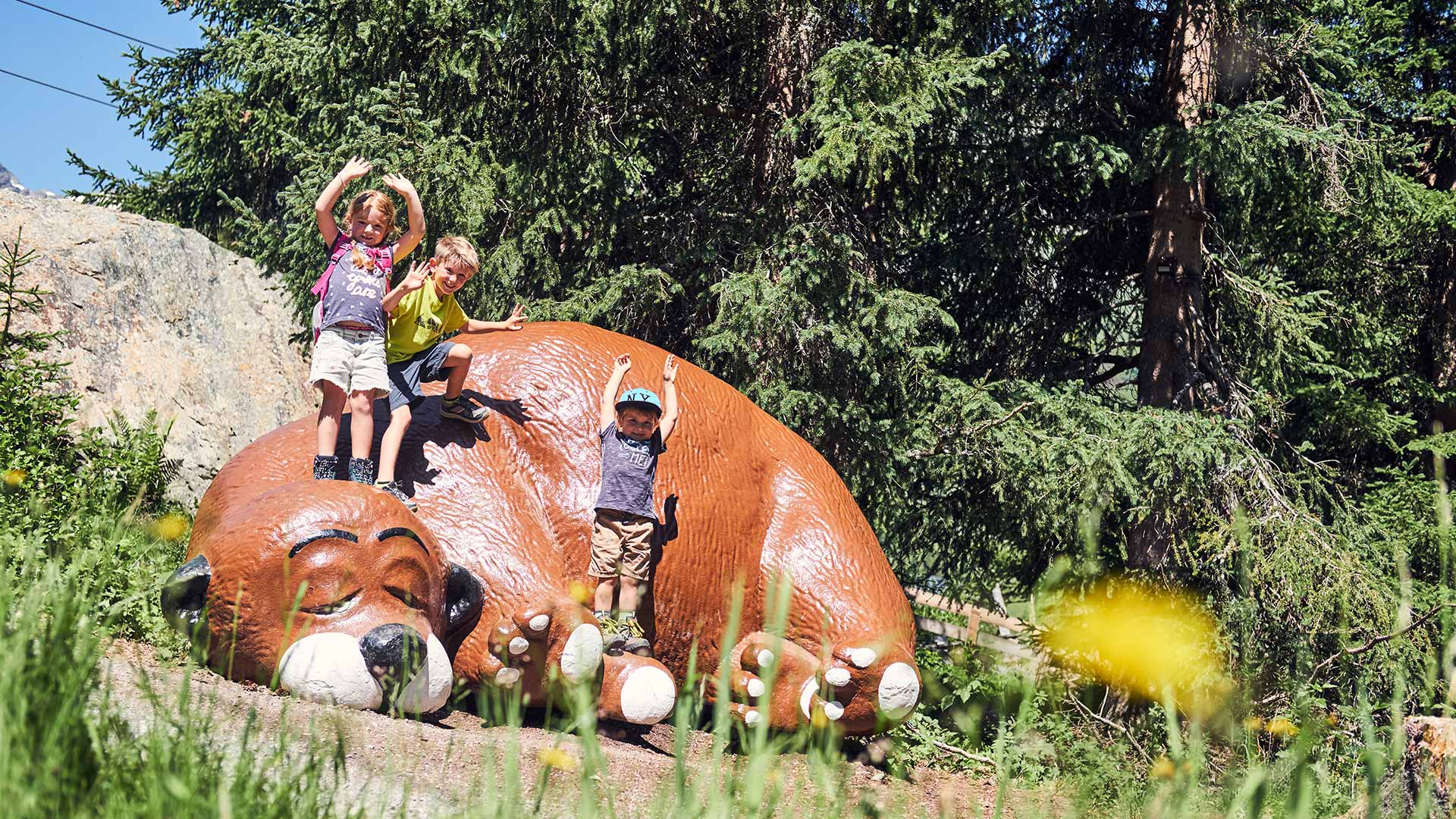 Drei Kinder klettern und posieren fröhlich auf einer riesigen Murmeltierfigur im Wald bei Sonnenschein.