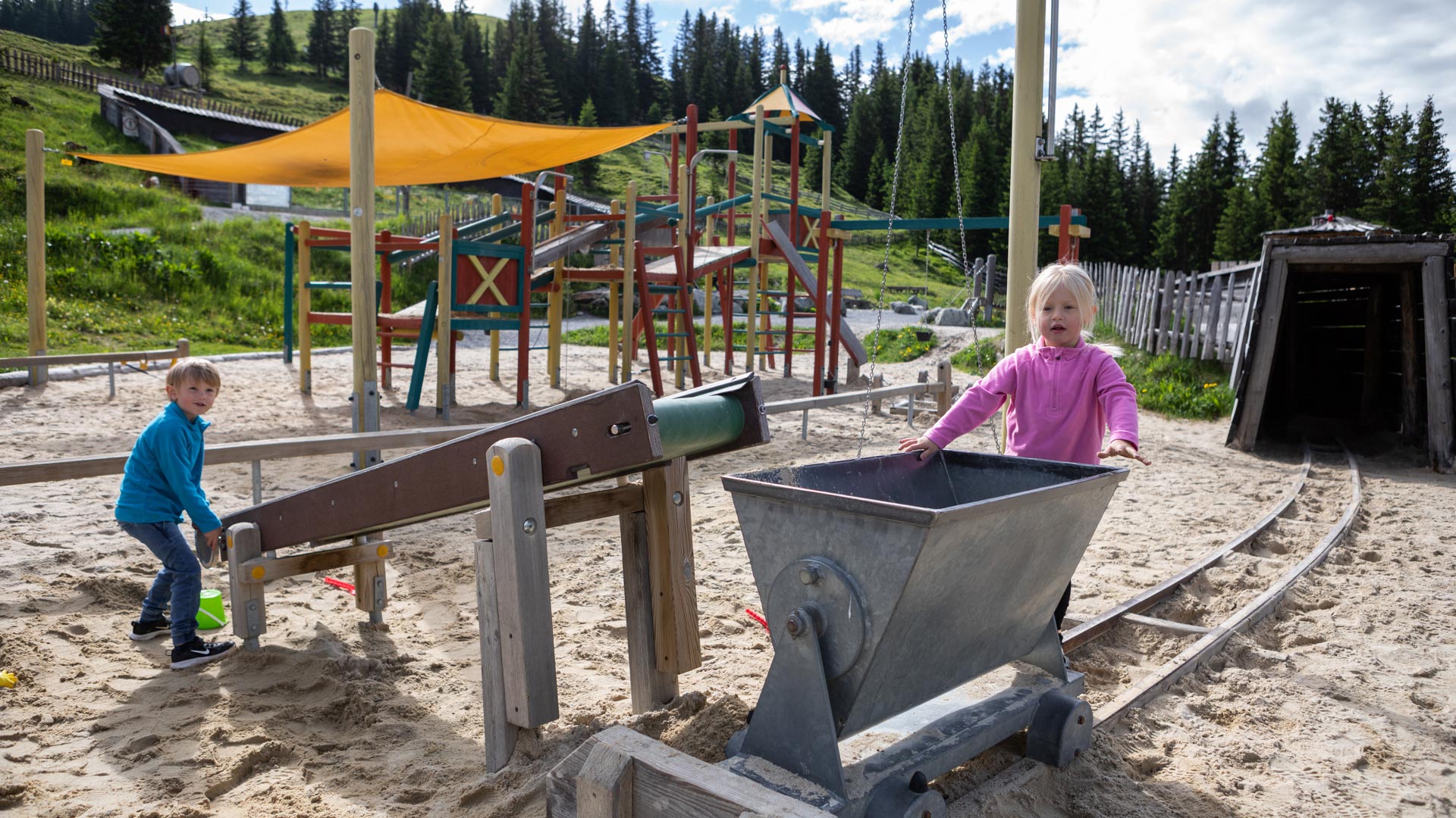 Zwei Kinder spielen auf einem Abenteuerspielplatz im Sand mit Wasserrinne und Lore vor einer Bergwaldkulisse.