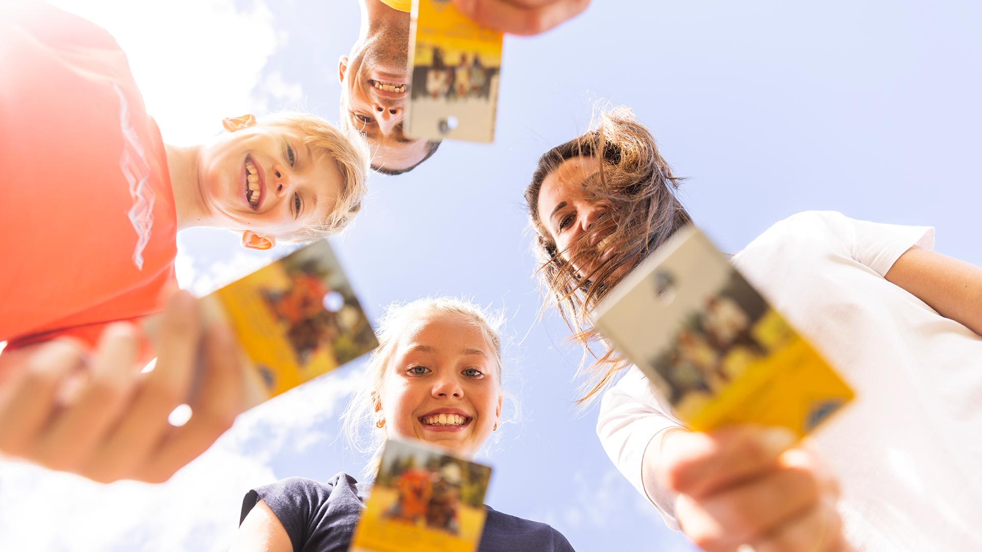 Vier fröhliche Menschen halten die Super.Sommer.Card in die Kamera und blicken lachend aus der Froschperspektive in den Himmel.