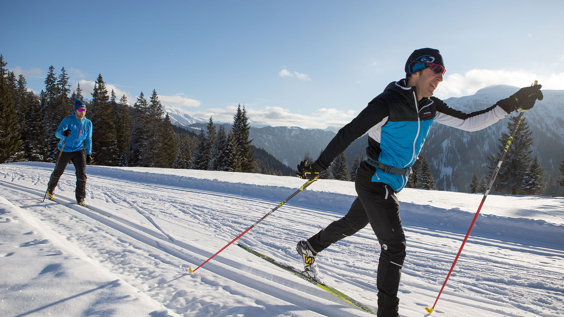 Zwei Langläufer gleiten sportlich durch verschneite Winterlandschaft mit Tannen und Bergblick bei Sonnenschein.